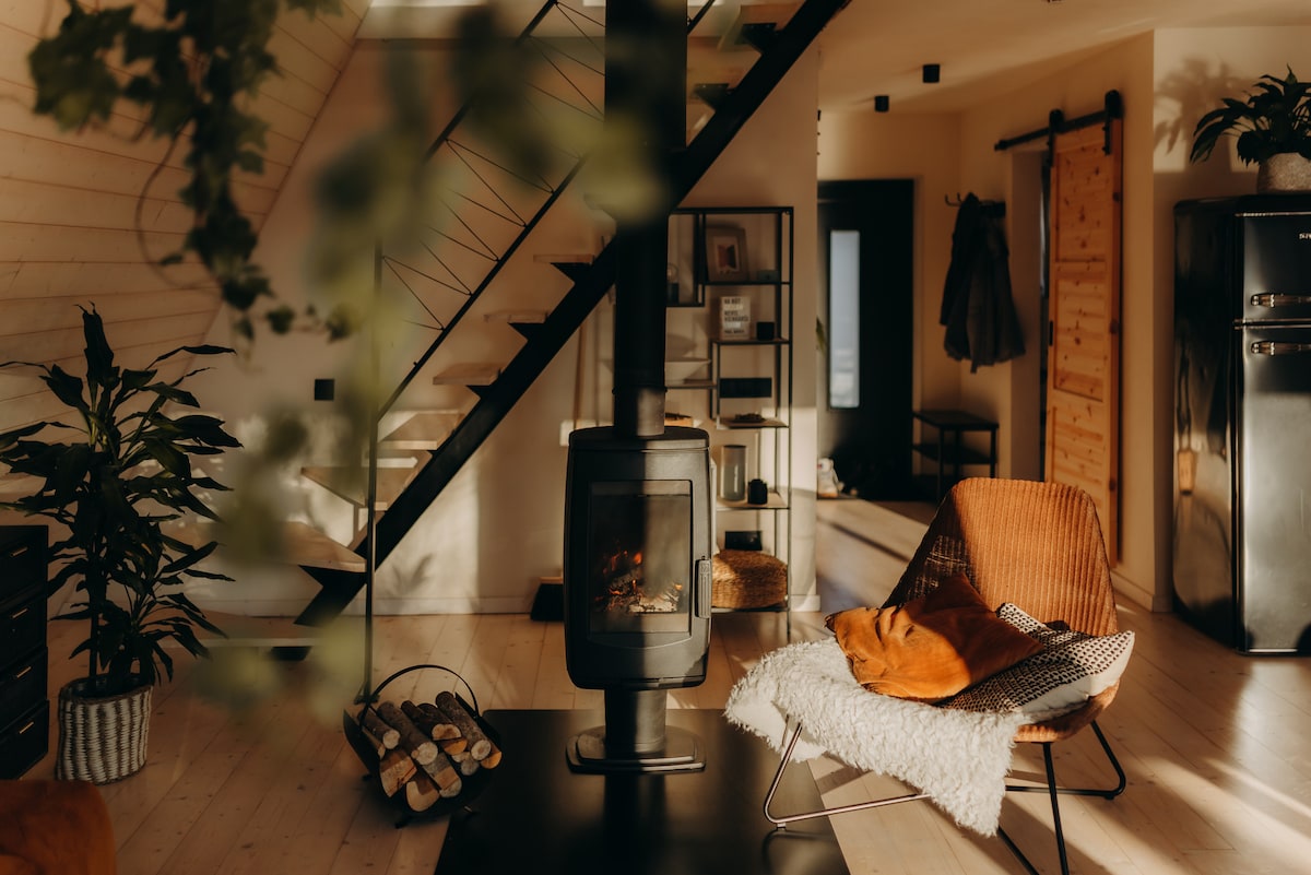 A modern living area is featured with a black wood-burning stove at its center. A woven chair is positioned nearby, adorned with a soft throw blanket. Natural light filters through large windows, highlighting the wooden staircase and fresh greenery.