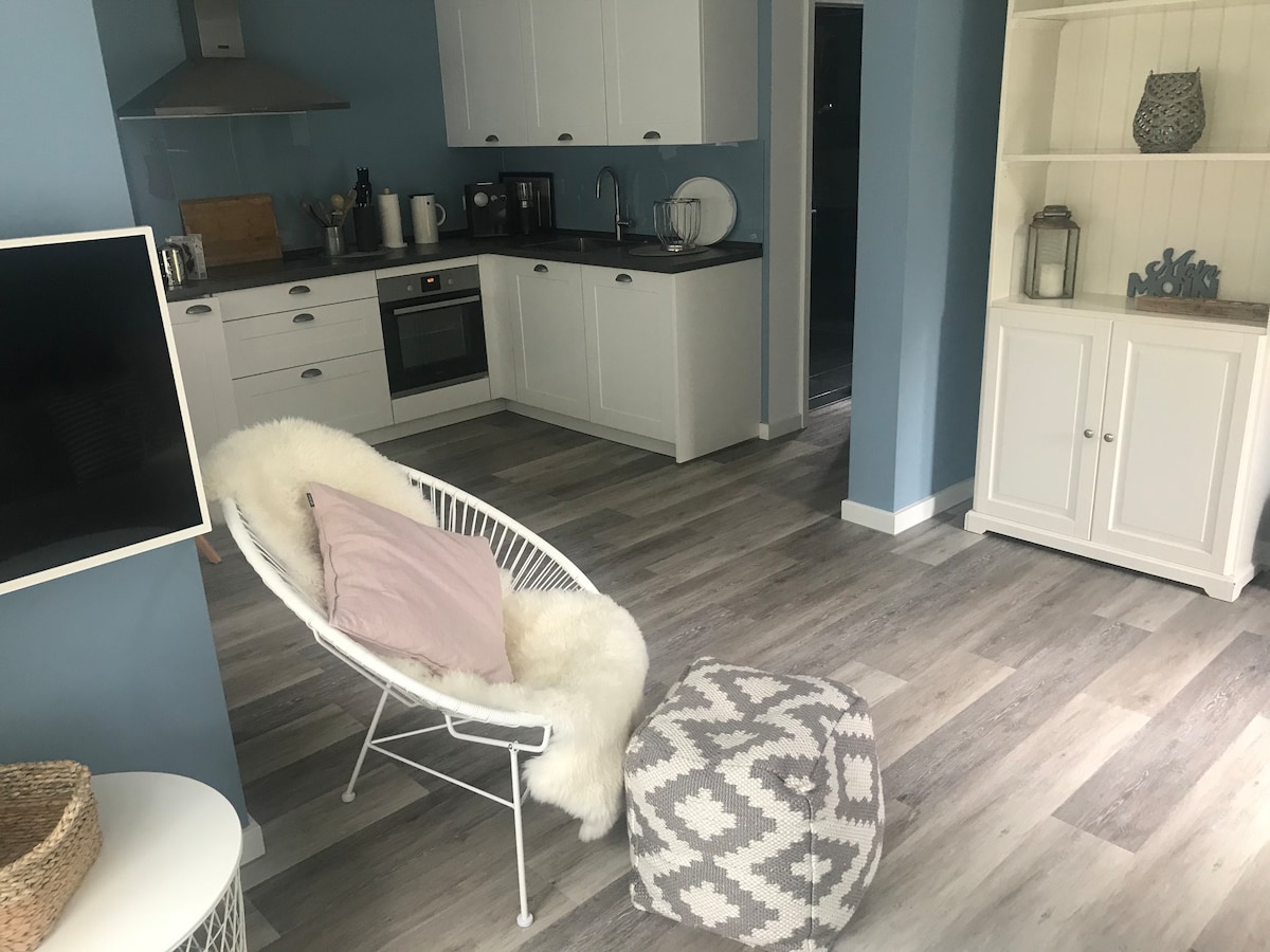 A bright living space features a cozy chair draped with a soft fur throw and a patterned pouf for added comfort. The modern kitchen, equipped with essential appliances, is visible in the background, complemented by light wood accents and a welcoming color palette.