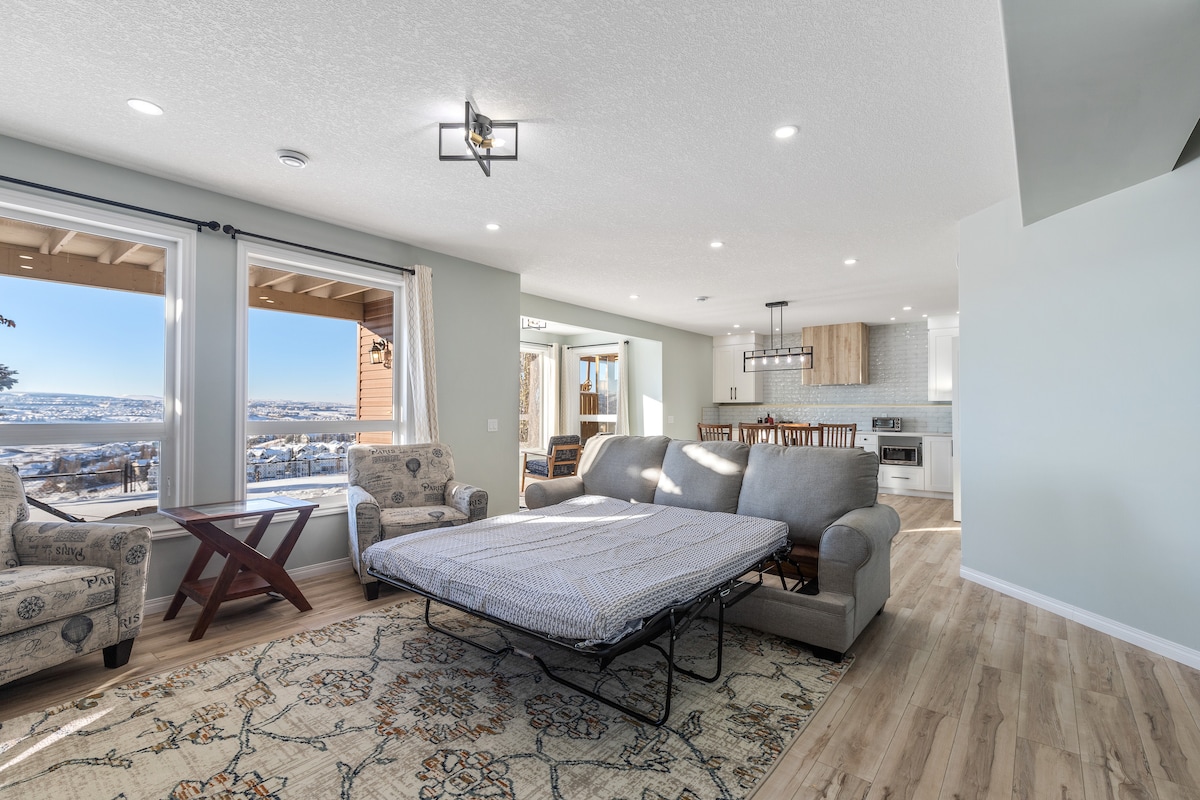A spacious living area features a comfortable sofa bed, with two accent chairs positioned nearby. Large windows allow natural light to fill the space, showcasing scenic views of the surroundings. A patterned area rug adds texture to the light-toned flooring, enhancing the inviting atmosphere.