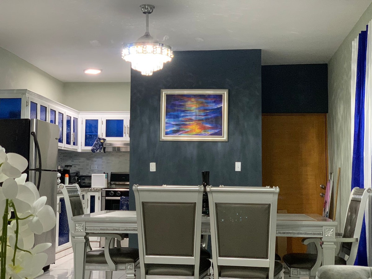 A dining area is illuminated by a chandelier, featuring a large table complemented by several chairs. A colorful piece of artwork adorns the wall, with a modern kitchen visible in the background that includes dark cabinets and appliances.