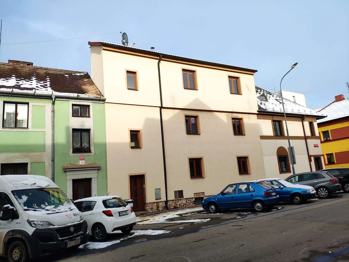 A three-story apartment building is shown, featuring a light-colored facade with brown trim. Windows are positioned symmetrically, casting shadows on the exterior. Vehicles are parked along the street in front, including a white van and a blue car, with nearby houses displaying vibrant colors.