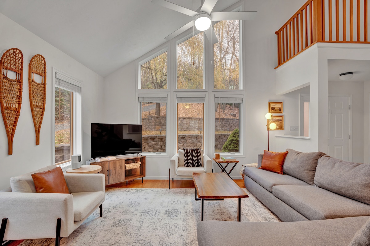 The living room features large windows that allow natural light to fill the space. A comfortable sectional sofa and armchair provide seating, complemented by a coffee table. A smart TV is positioned on a media console, with snowshoe decor adorning the walls.