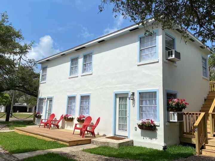 The exterior of a two-story white building is shown, featuring large windows framed in blue. A wooden deck with red chairs invites relaxation, while flower boxes with blooming plants add color. Stairs lead to the second level, where additional units are accessible.