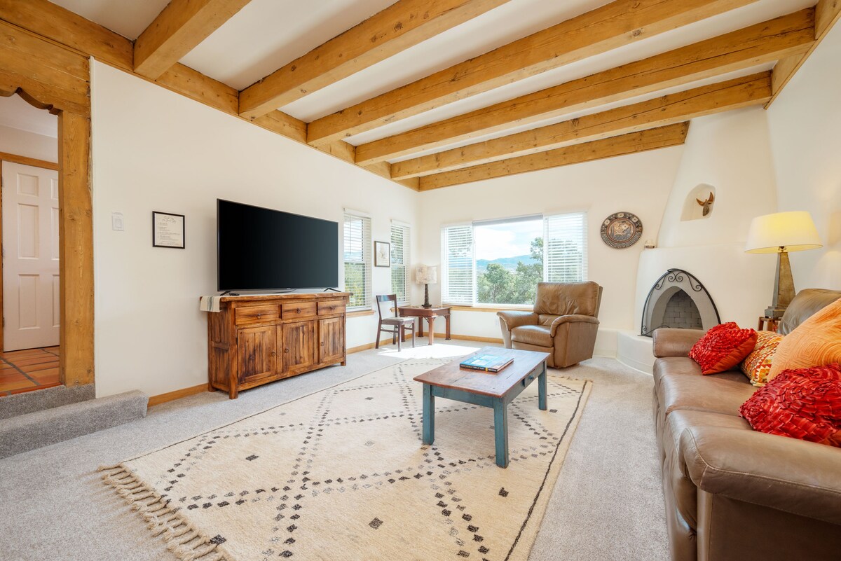 A spacious living room features high wooden beams overhead and soft carpeting. A large smart TV is mounted on the wall, opposite a comfortable leather sofa adorned with colorful cushions. Natural light streams through a large window, illuminating a cozy reading nook with an armchair.