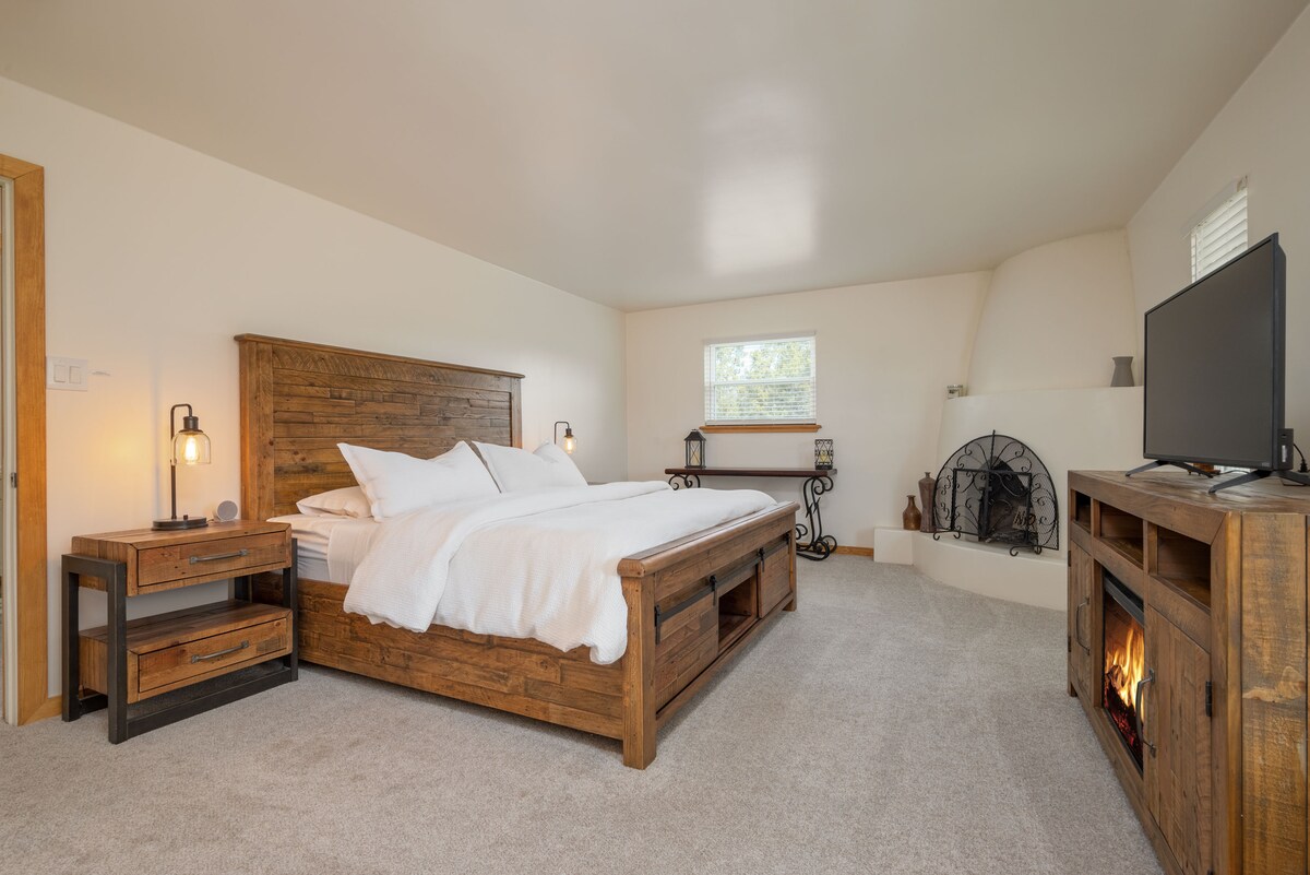 A spacious bedroom features a wooden bed frame with fluffy white bedding. A cozy electric fireplace is positioned in a corner, complemented by a decorative fire screen. A nightstand with a lamp is by the bed, and a small window allows soft natural light to enter.