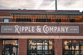 The exterior of a brick building displays the sign for Ripple & Company, showcasing the establishment's name in stylish lettering. Warm string lights are arranged above the entrance, hinting at a lively atmosphere. Large windows provide glimpses of the interior.