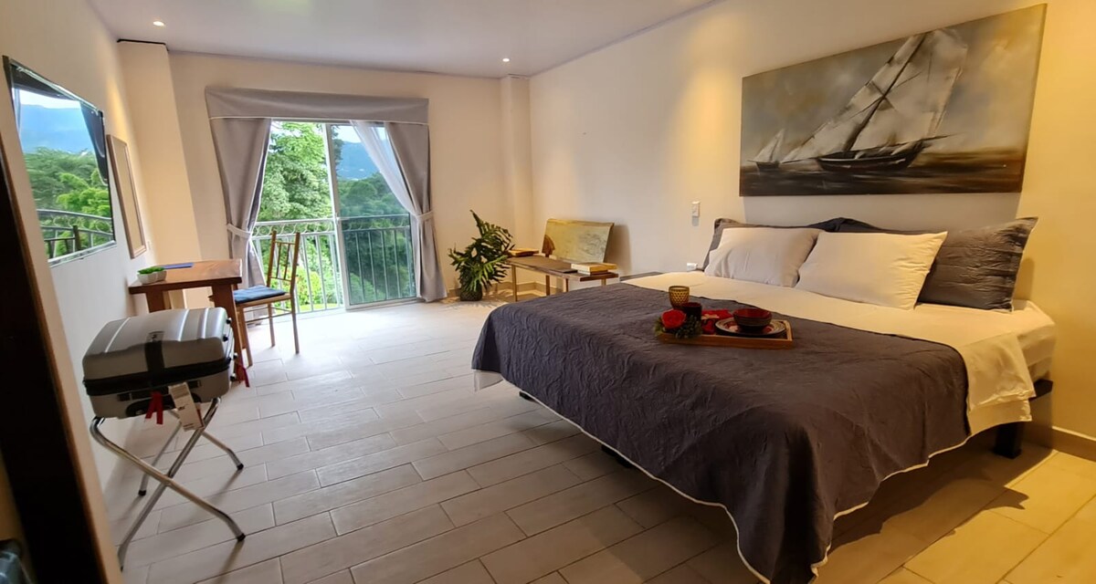A spacious room features a large bed with a neutral-colored blanket, complemented by a simple art piece depicting a sailboat. Natural light filters through the balcony window, which opens to a lush outdoor view. A small seating area and a foldable table are also present.