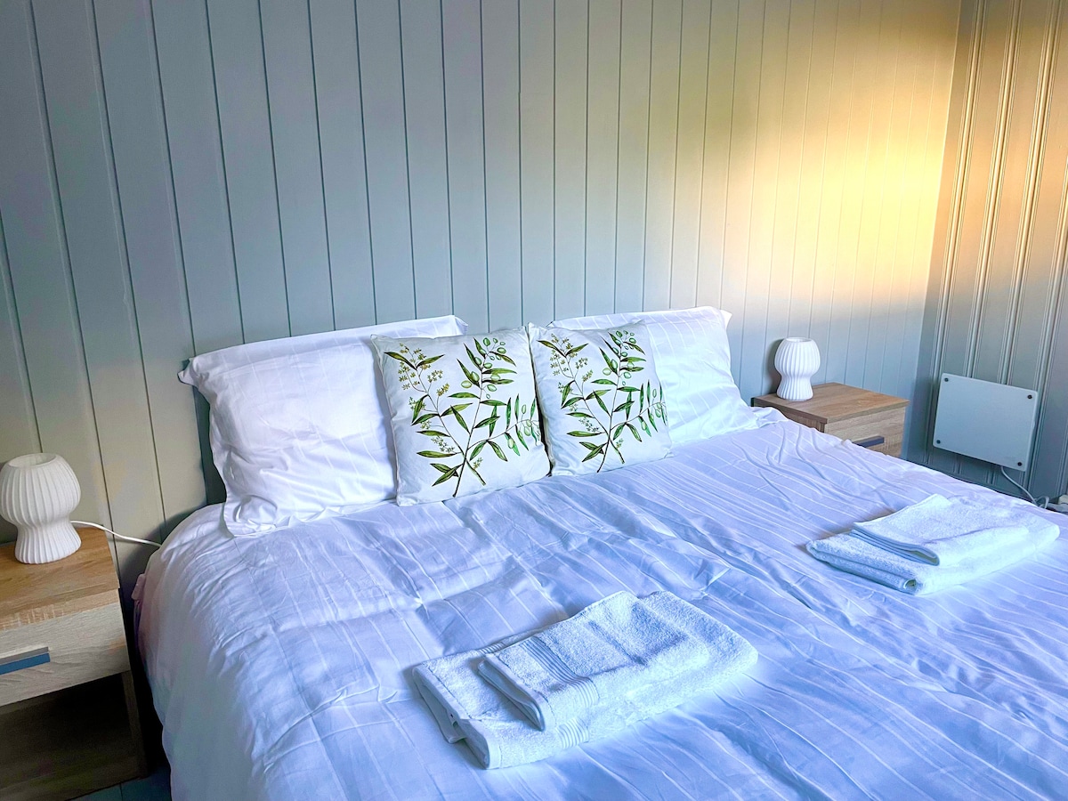 A spacious bed is dressed with crisp white linens, centered between two nightstands with soft, textured lamps. An embroidered pillow adds a touch of nature-inspired design. Fresh towels are neatly arranged at the foot of the bed, welcoming guests for a comfortable stay.