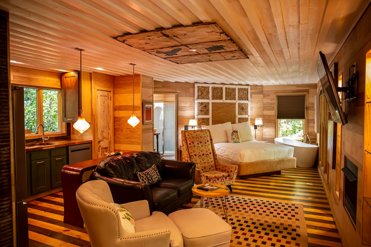 The main room features custom wood paneling with a loft-like feel, illuminated by pendant lights. A spacious bed is positioned beside a soaking tub, with a cozy living area containing an accent chair and a small side table. Windows offer views of the surrounding woods.