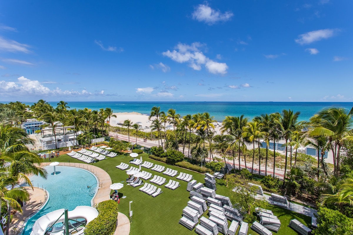 The image captures a vibrant view of a beachside area featuring a large pool surrounded by sun loungers under palm trees. The expansive ocean stretches into the horizon, complemented by a clear blue sky with scattered clouds. Green landscaping adds an inviting touch to the scene.