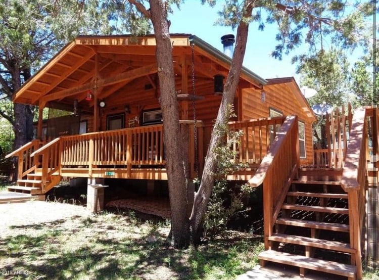 The cabin exterior features a wooden façade with a spacious wrap-around deck. Stairs lead up to the entrance, while a tree provides shade nearby. A swing hangs from a branch, enhancing the outdoor setting surrounded by greenery.