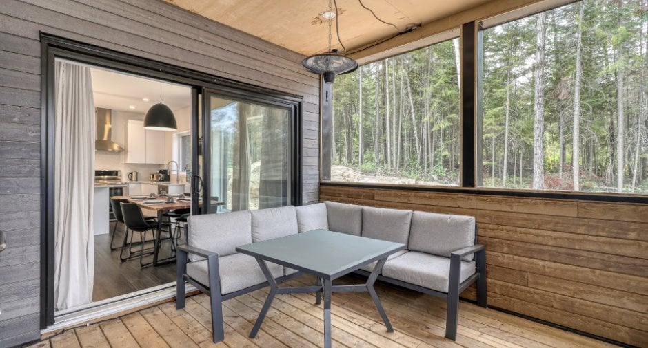 A covered terrace is furnished with a sectional sofa and a glass-top table. Large windows offer views of the surrounding forest, while a door leads to the indoor living space. The wooden flooring adds a natural touch to the setting.