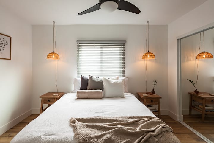 Სტუმრების საძინებელი Rattan გულსაკიდი განათებით, ორადგილიანი საწოლით და ცოცხალი ZZ მცენარეების ჭრით.