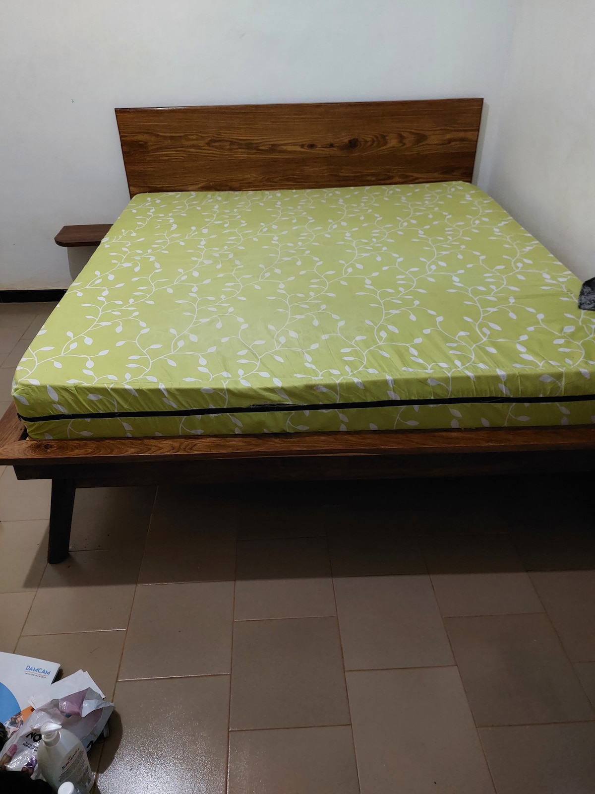 A spacious bed is set against a simple wooden headboard, featuring a light green mattress cover adorned with a white floral pattern. The room is minimally furnished with neutral-colored tiles, contributing to a calm and uncluttered environment.