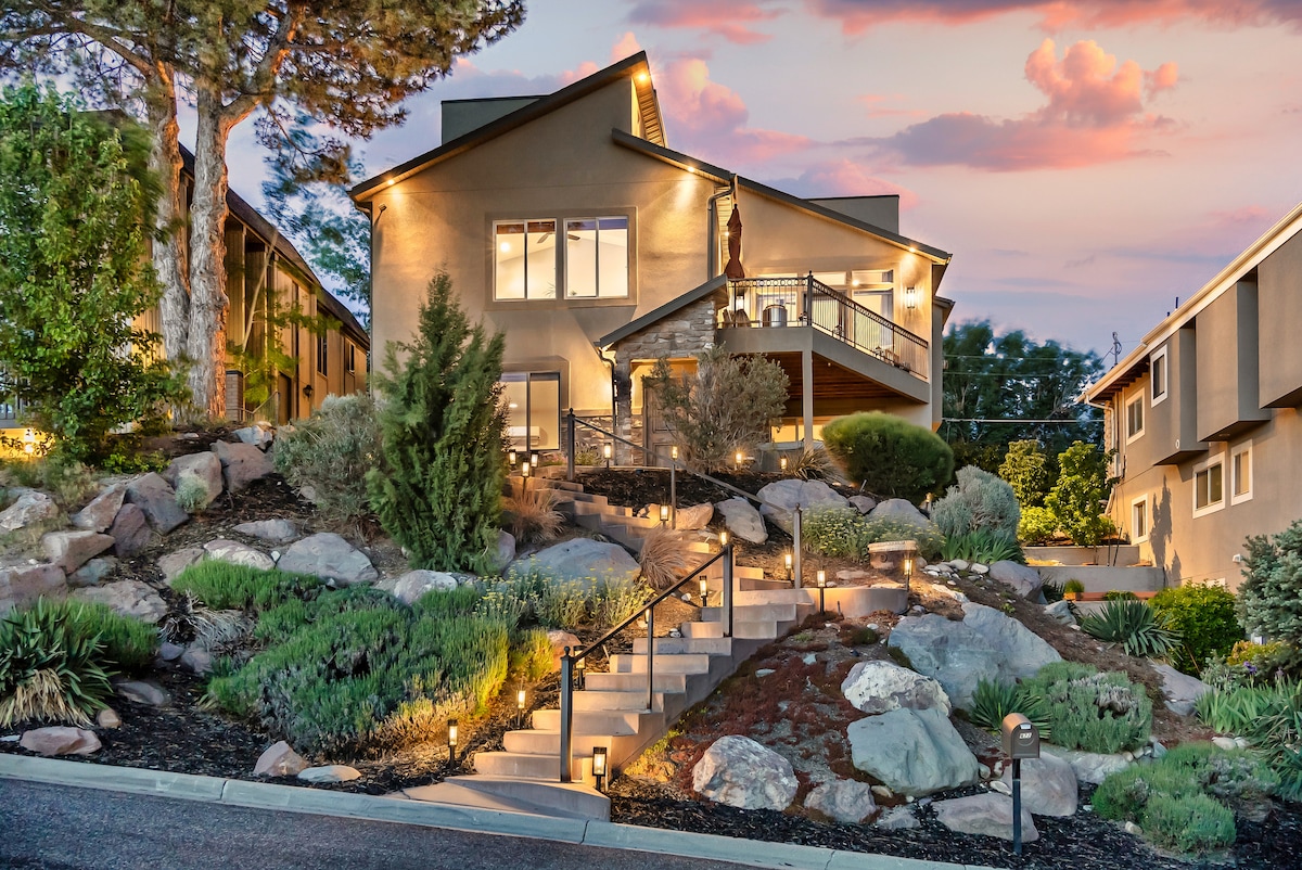 An inviting exterior showcases a modern two-story home surrounded by carefully landscaped greenery and rocks. Soft outdoor lighting highlights the pathway leading up to the entry, with a dramatic sunset illuminating the sky in vibrant hues.