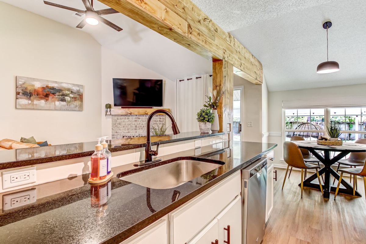 A well-appointed kitchen features dark granite countertops, showcasing a stainless-steel sink and dishwasher. The space is complemented by wooden beams and a cozy dining area with a round table, surrounded by four stylish chairs, with natural light streaming through the nearby windows.