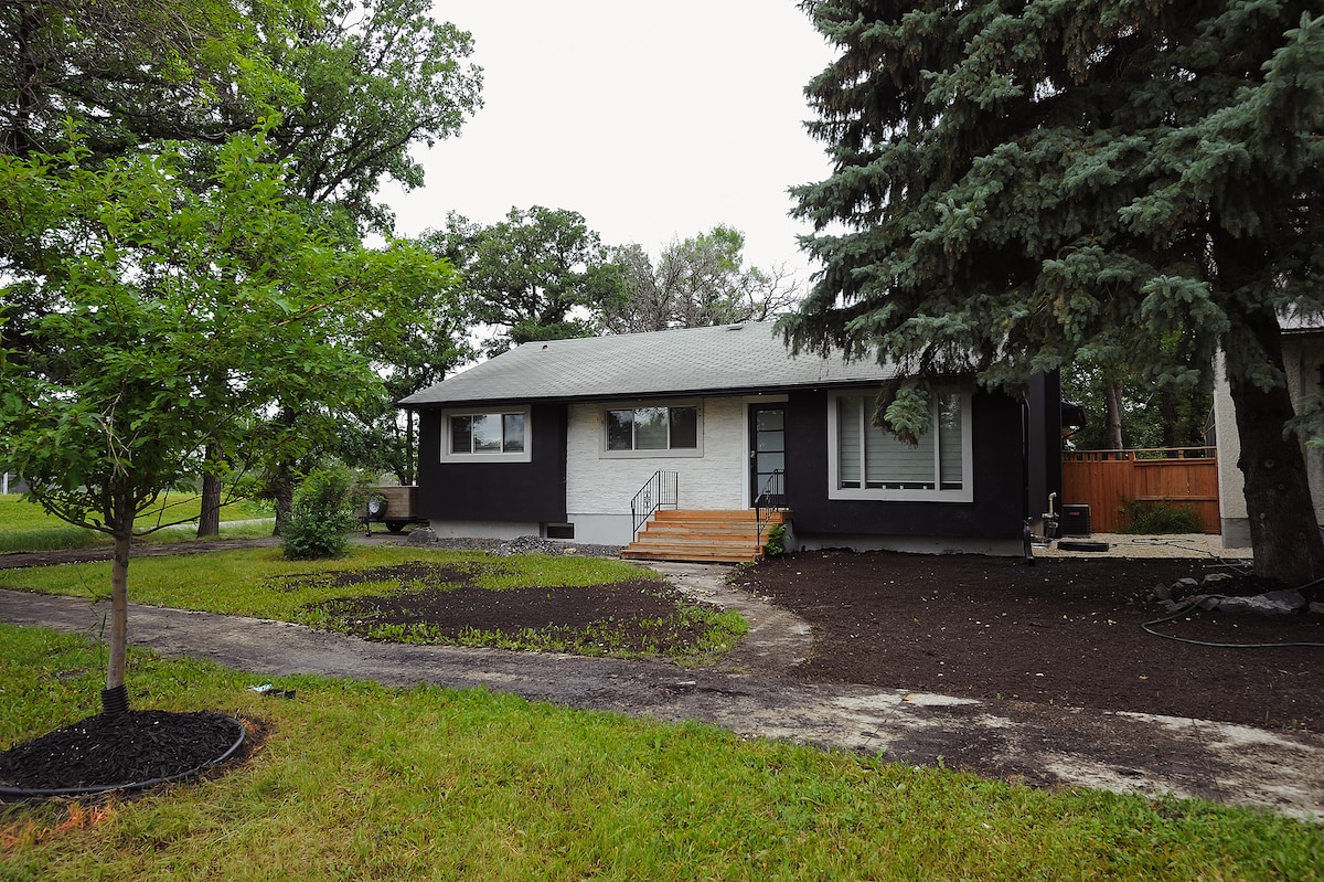 The exterior of a bungalow is shown, featuring a mix of dark siding and light stone accents. A pathway leads to the entrance, which is flanked by large windows. Surrounding the home are trees and a well-maintained lawn, enhancing the inviting appearance of the property.