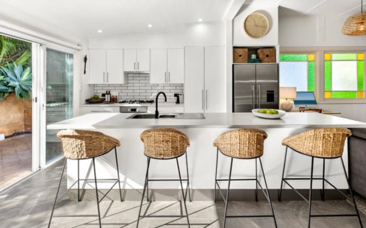 The modern kitchen features a spacious island with four woven bar stools positioned around it. Bright white cabinetry is complemented by a sleek countertop. Natural light enters through sliding glass doors and a stained glass window adds a pop of color.