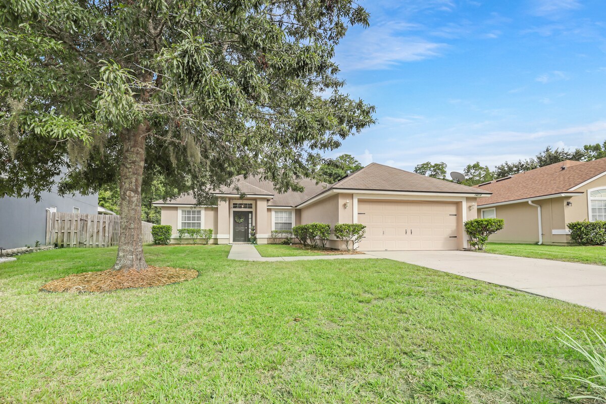 The exterior of the home features a neatly manicured front yard with lush green grass and a large tree providing shade. A wide driveway leads to a two-car garage, and the entryway is framed by decorative landscaping, creating an inviting approach.