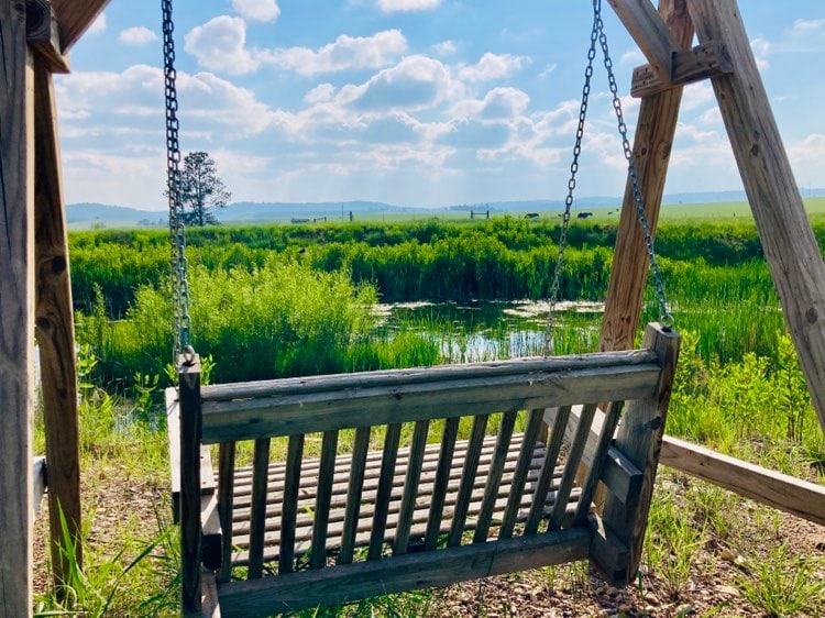 A wooden swing is suspended from a sturdy frame, facing a serene view of lush greenery and a calm body of water. The sky displays soft, fluffy clouds above, providing a tranquil backdrop for relaxing moments in nature.