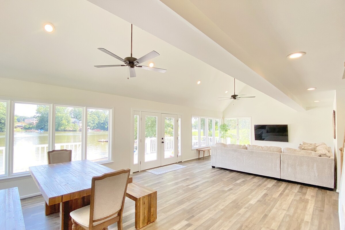 A spacious living area features large windows that provide views of the lake. Natural light fills the room, complemented by ceiling fans. A wooden dining table is positioned next to a comfortable sectional sofa, with a wall-mounted TV visible in the background.