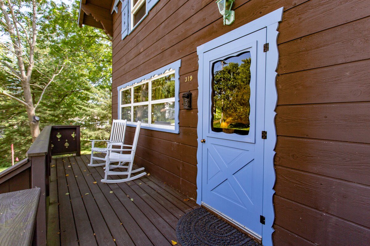 The entryway features a light blue door with a glass panel, surrounded by rich brown wooden siding. A welcoming porch is adorned with two white rocking chairs, resting on a wooden floor lined with leaves from nearby trees.