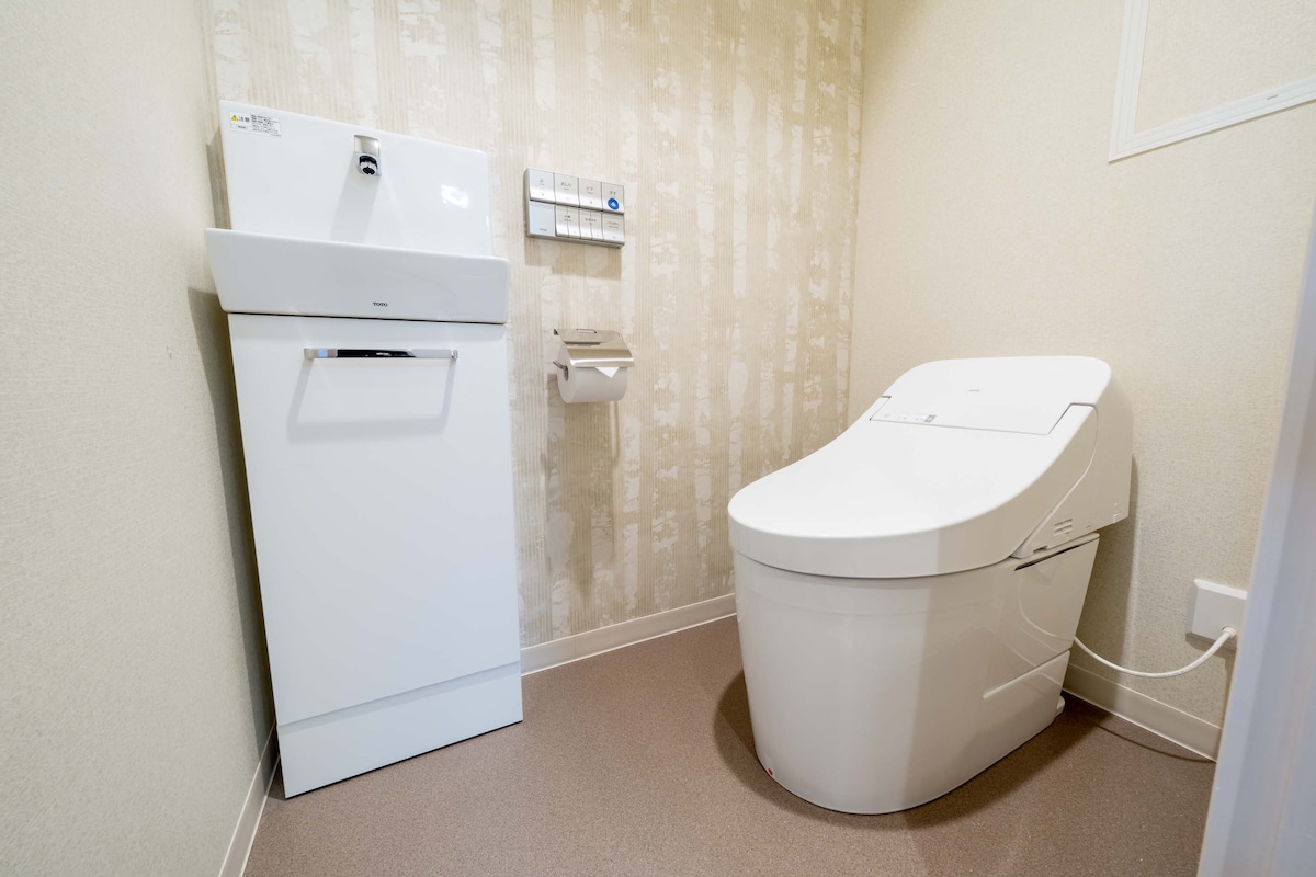 The bathroom is equipped with a modern toilet featuring additional controls. A sink and faucet are positioned nearby, providing convenience. The walls display a subtle textured pattern, and the floor is finished in a smooth surface, contributing to a clean and functional atmosphere.