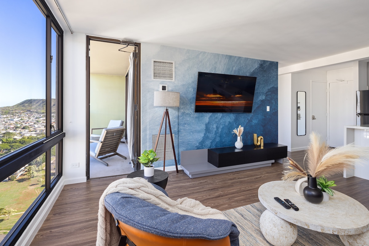 A modern living area features a large TV mounted on a blue-accented wall. A comfortable armchair and round coffee table are arranged on warm wood flooring. An open lanai access point reveals views of the surrounding landscape, while a stylish plant adds a touch of greenery.