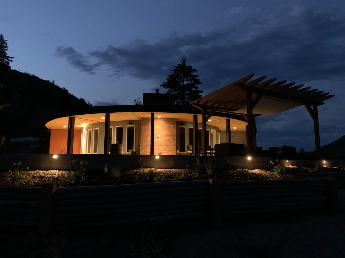The roundhouse is illuminated under a twilight sky, showcasing its curved architecture. Outdoor seating is located under a wooden pergola, with low-profile lighting highlighting the landscaped area. The surrounding natural landscape is framed by trees and distant hills, enhancing the tranquil evening atmosphere.