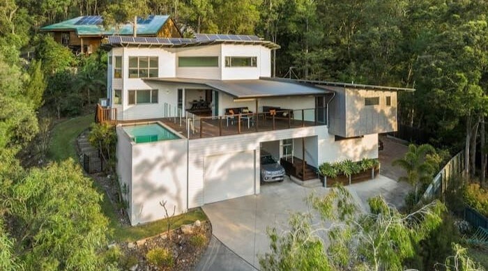 The modern three-level home is nestled among trees, featuring a spacious front deck with a sparkling pool. Large windows allow natural light to fill the interior. A driveway leads to the carport, positioned alongside the house, amidst a serene natural landscape.