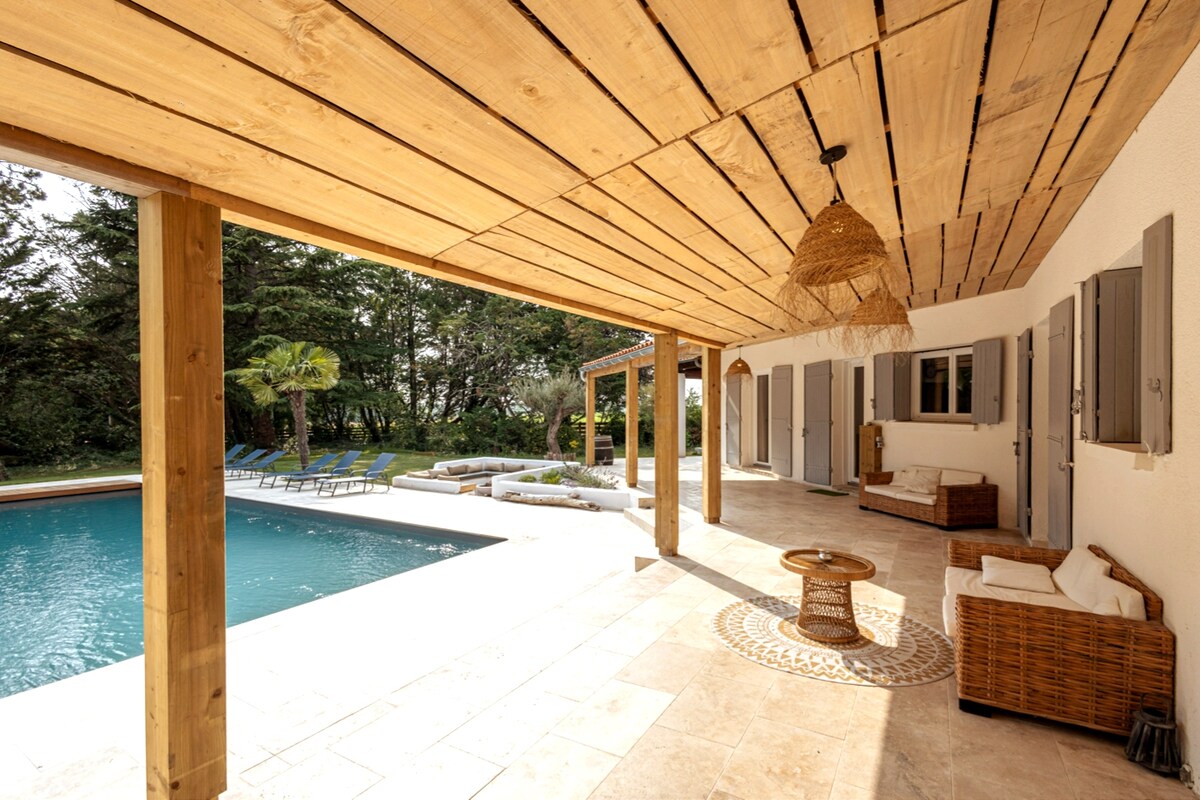 An outdoor terrace features a wooden ceiling and natural wicker furniture, providing a comfortable seating area. A pool is visible in the background, surrounded by lounge chairs and a landscaped garden, creating a serene space for relaxation.