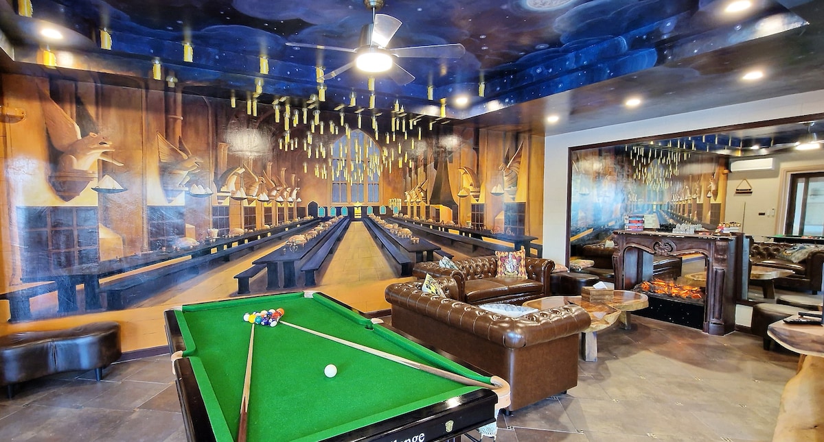 A spacious game room is showcased, featuring a green snooker pool table in the foreground. The walls are adorned with a mural depicting a grand hall, illuminated by hanging lights. Cozy seating areas are visible, enhancing the room's welcoming ambiance.