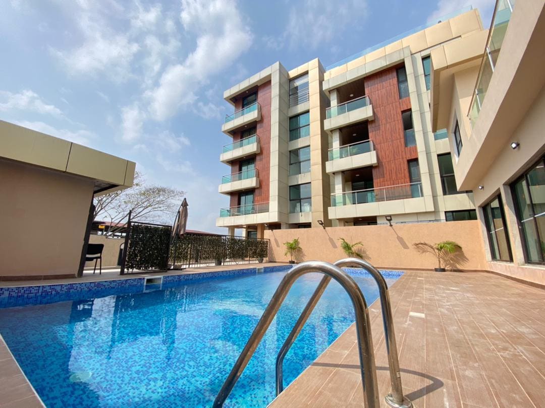 A modern building stands in the background, featuring multiple balconies with wooden accents. In the foreground, a shimmering pool is framed by a tiled deck, with lounge chairs and planters adding to the serene environment. Blue sky and clouds enhance the inviting atmosphere.