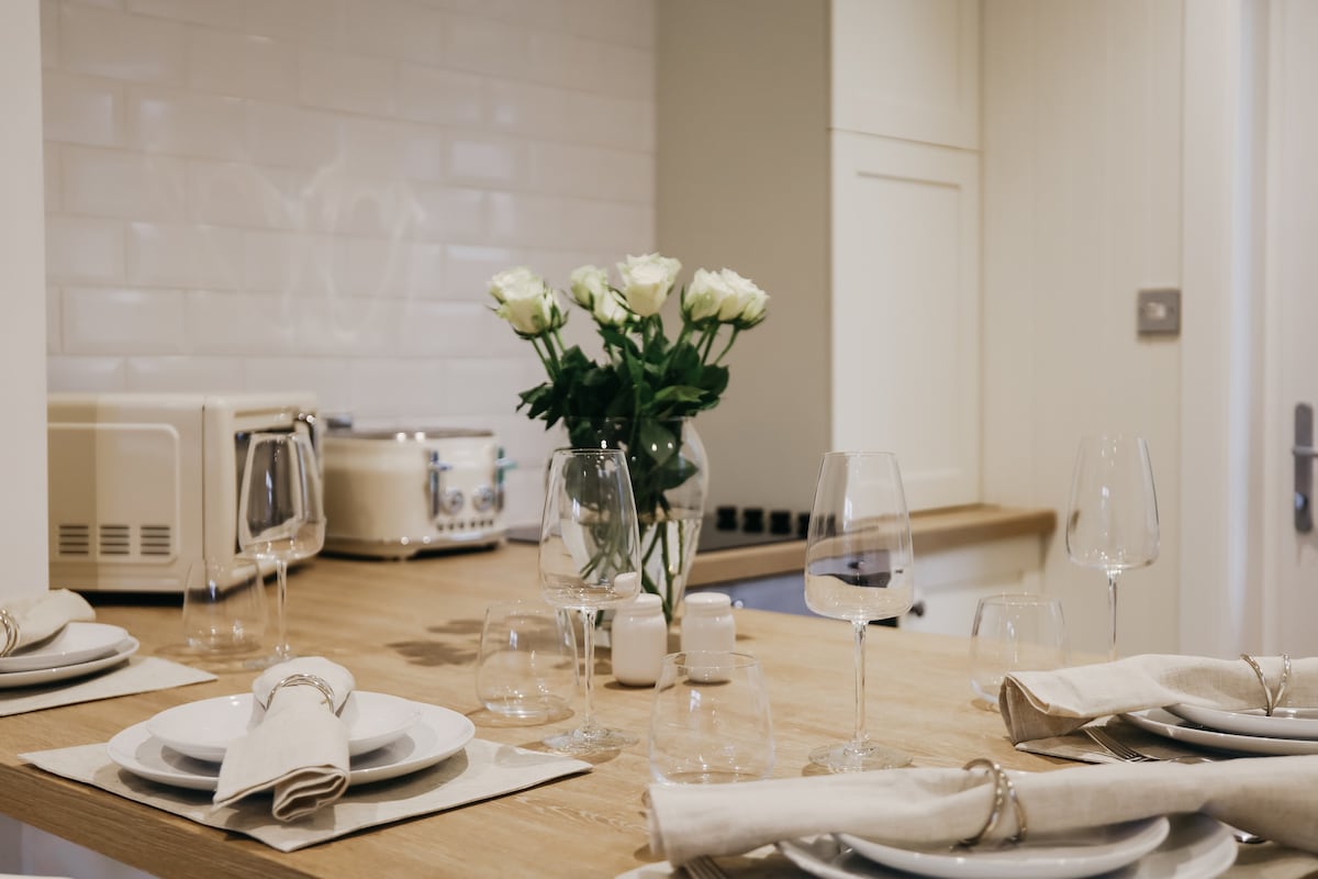 A dining table is set with plates and glassware, accompanied by folded napkins. A vase of fresh roses is placed at the center, adding a touch of elegance. A beige countertop and a retro appliance are visible in the background, contributing to a modern yet inviting kitchen environment.