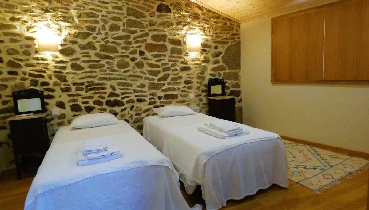 The sleeping area features two twin beds draped in white linens. Each bed is flanked by wooden nightstands with vintage lamps. The stone wall adds a rustic charm, while a light wooden ceiling and a colorful area rug enhance the cozy atmosphere.