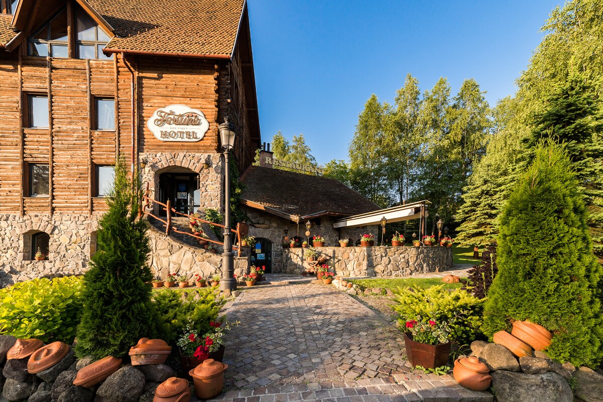 The exterior of the Eco-Boutique Hotel features rustic wooden architecture complemented by stone accents. A cobblestone path leads to a welcoming entrance adorned with potted plants and vibrant flowers, surrounded by lush greenery under a clear blue sky.