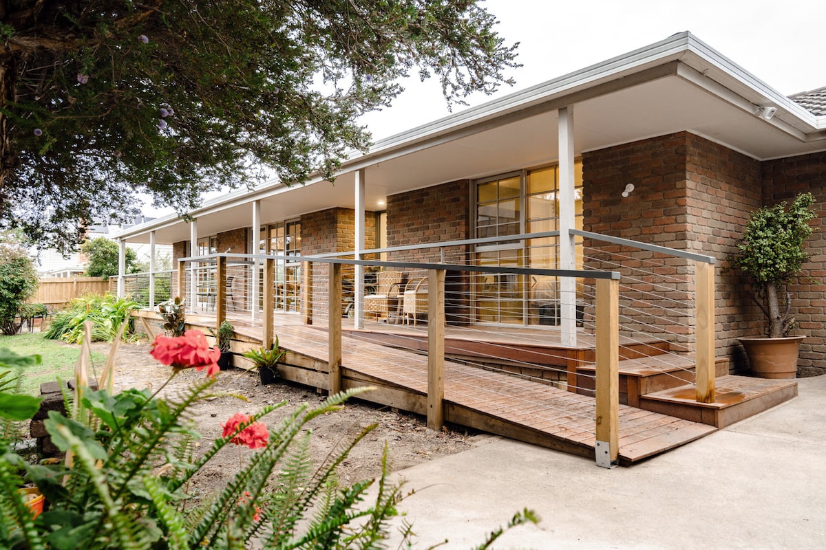 A spacious outdoor deck is highlighted, featuring a wooden ramp leading to the entrance. The area is surrounded by lush greenery, including blooming flowers and neatly arranged garden beds. Large windows reflect natural light, creating an inviting connection between the indoor and outdoor spaces.