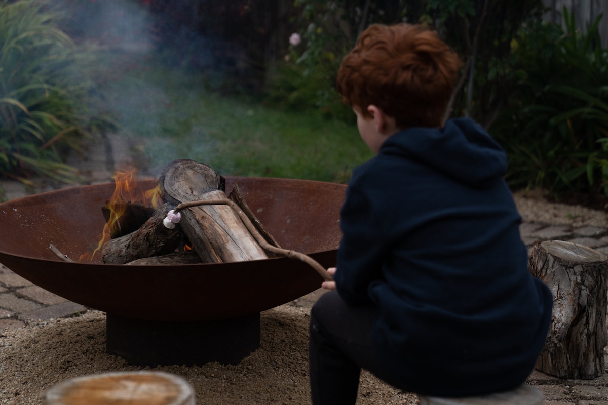 A fire pit is featured in a secluded corner of a garden, surrounded by gravel and logs. Flames dance atop a few logs, and smoke gently rises into the air. The scene captures a serene moment, inviting relaxation and storytelling.