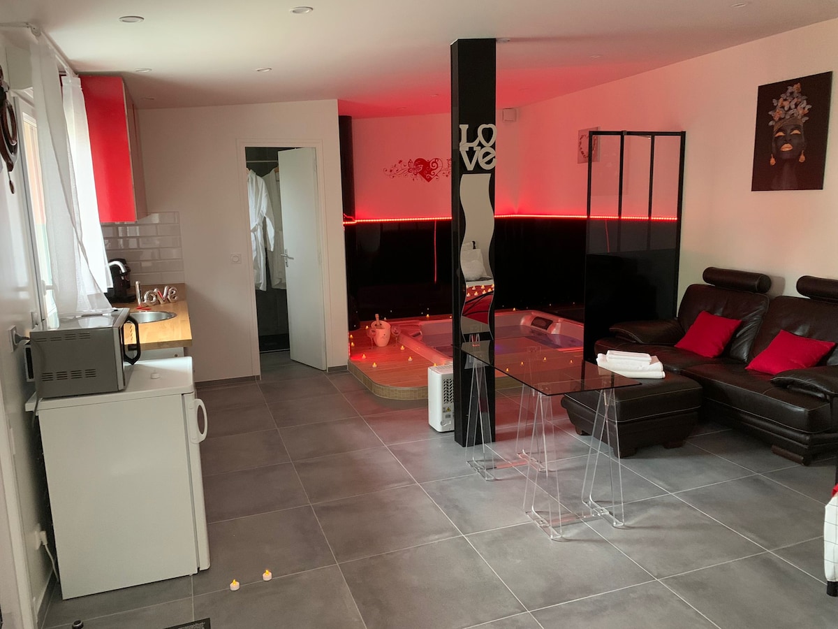 An interior space is presented featuring a comfortable seating area with dark leather sofas and ambient lighting. A compact kitchenette is situated in one corner, and a jacuzzi is centrally located, surrounded by candlelight, creating an intimate atmosphere. The walls display modern art.
