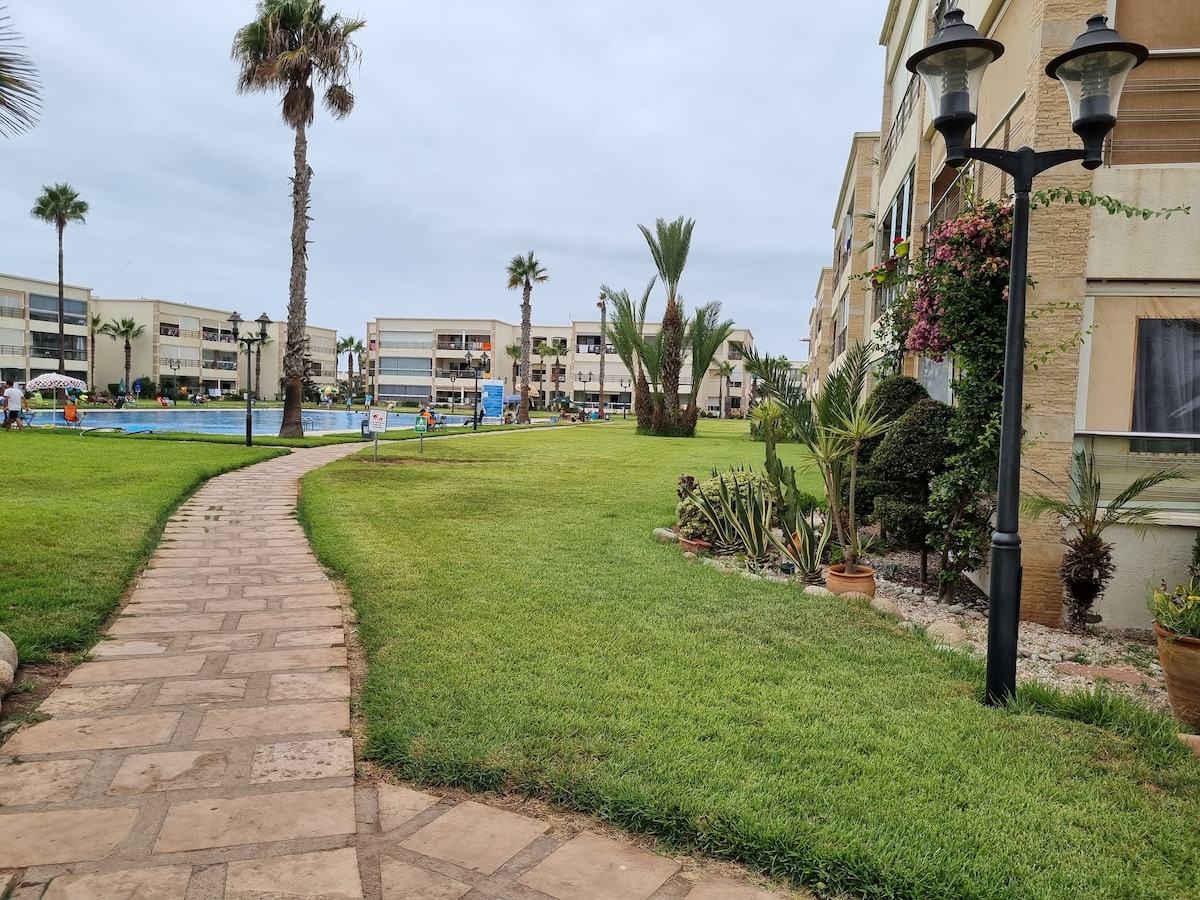 A stone pathway winds through a lush green lawn, bordered by palm trees and tropical plants. Soft lighting from a decorative lamp enhances the garden area adjacent to the buildings. A swimming pool is visible in the background, suggesting a tranquil outdoor space.