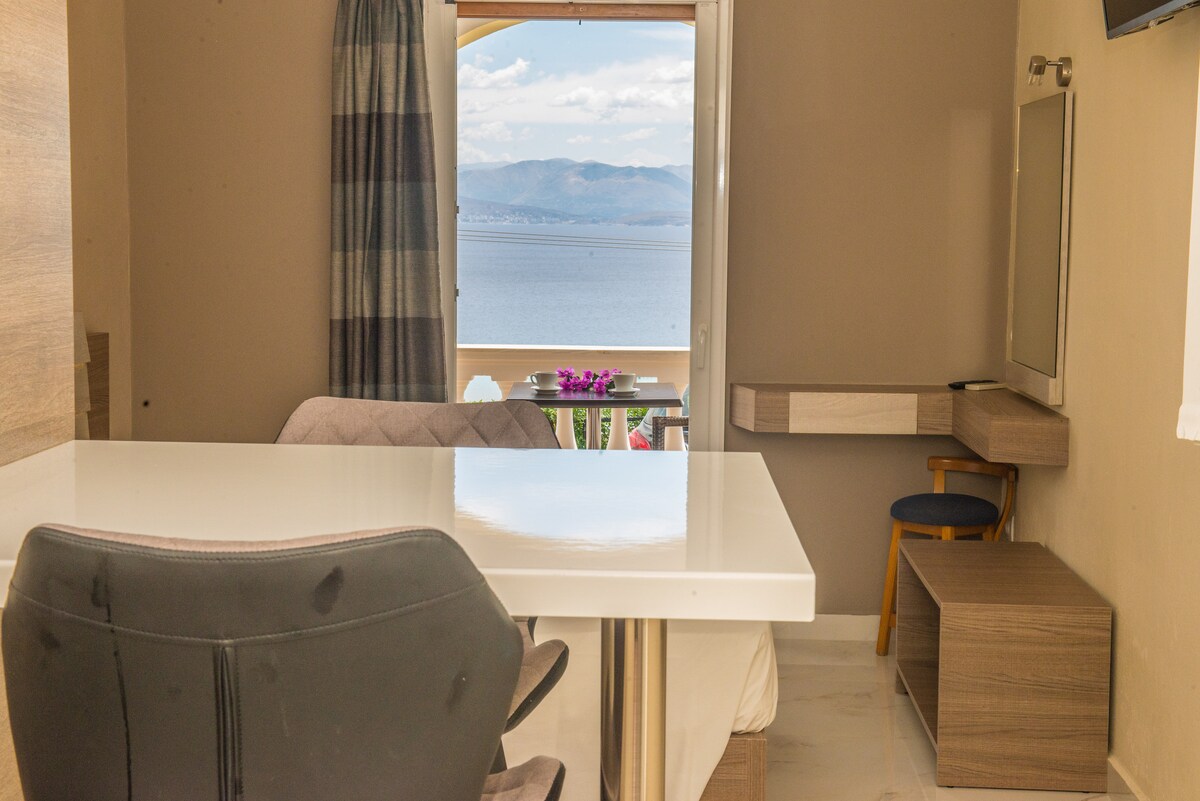 A modern workspace is positioned next to a large window, offering views of the sea and mountains. A comfortable chair rests at the desk, with a small mirror mounted above. Soft natural light fills the area through the partially opened curtain.