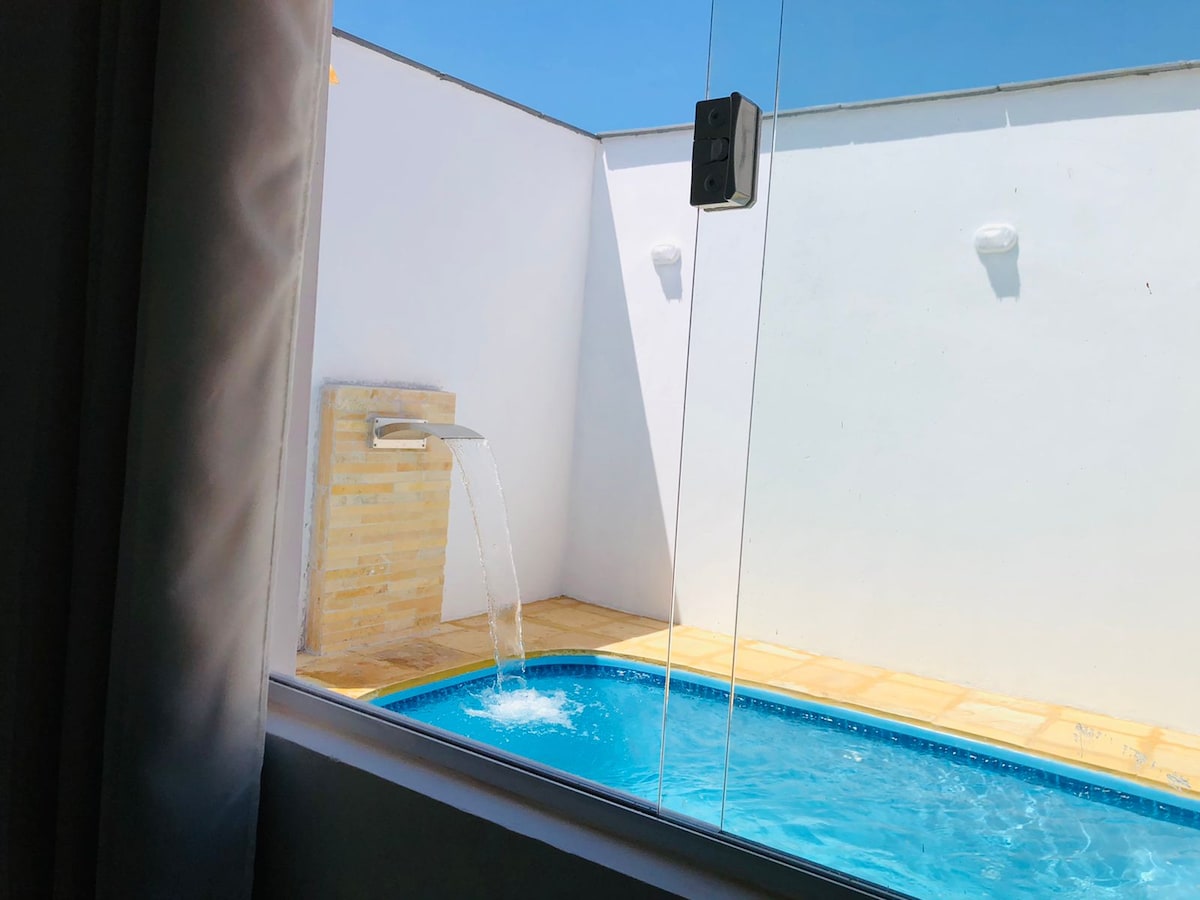 A private pool can be seen through a large glass window, featuring a wall-mounted water feature that gently pours into the water. Sunlight reflects off the clear blue surface of the pool, surrounded by a tiled deck and white walls.