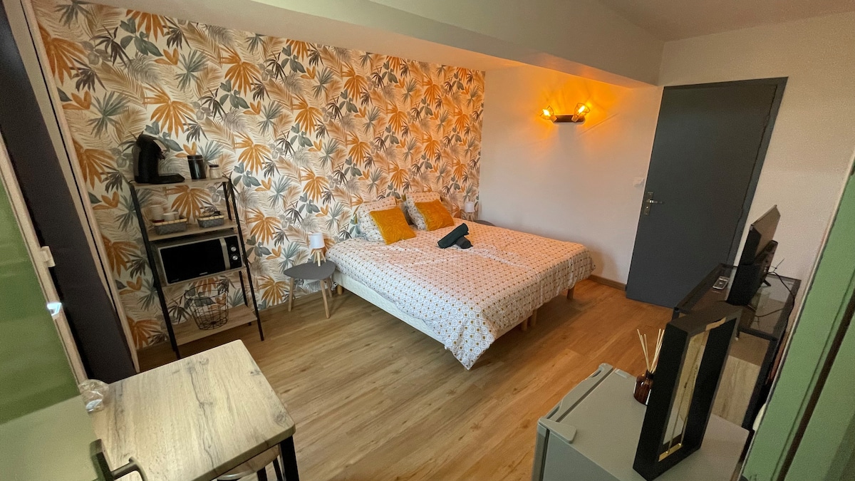A modern room showcases a spacious bed with decorative throws against a floral patterned accent wall. A small kitchenette is visible, along with a table and two chairs. The floor is covered in light wood, contributing to a fresh and contemporary look.