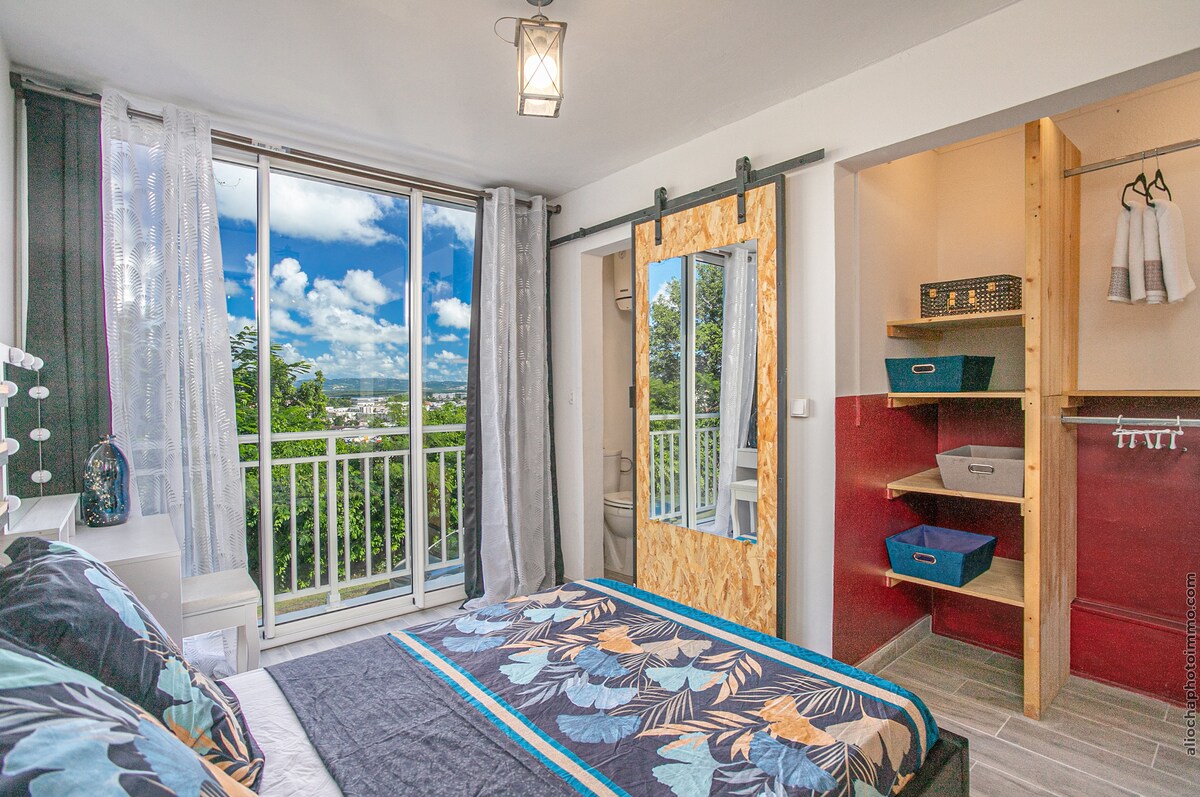 A well-organized bedroom showcases a spacious bed adorned with a patterned blanket. Large windows allow natural light to flood the room, revealing scenic views of the surroundings. A full-length mirror and a closet with storage options are visible, enhancing the room's functionality.