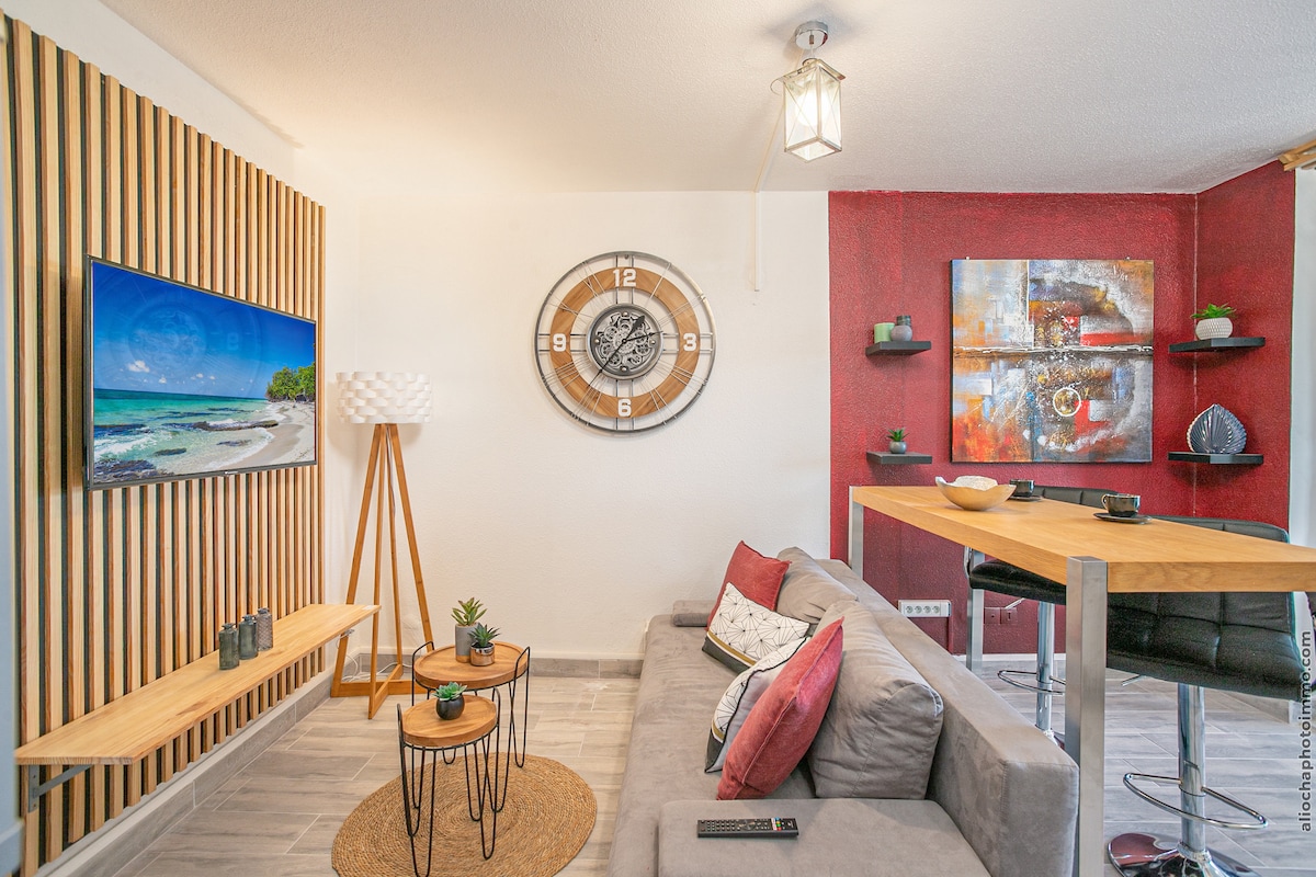 A cozy seating area is presented with a gray sofa adorned with red and white cushions. A sleek wooden table stands nearby, complemented by decorative plants. On the wall, a round clock and a colorful artwork add visual interest, while a TV is mounted opposite the seating.