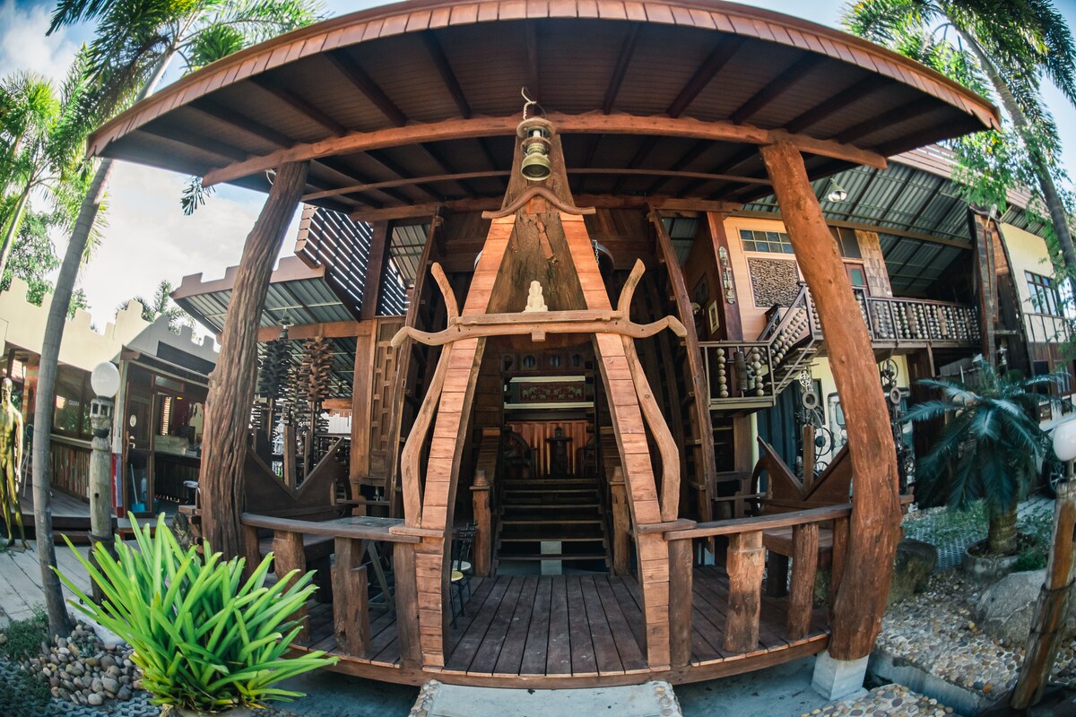 A wooden structure showcases intricate design elements and rustic charm. The open-air porch features uniquely carved seating areas and is surrounded by lush greenery. Natural light filters through the trees, creating an inviting space that blends seamlessly with the outdoor surroundings.