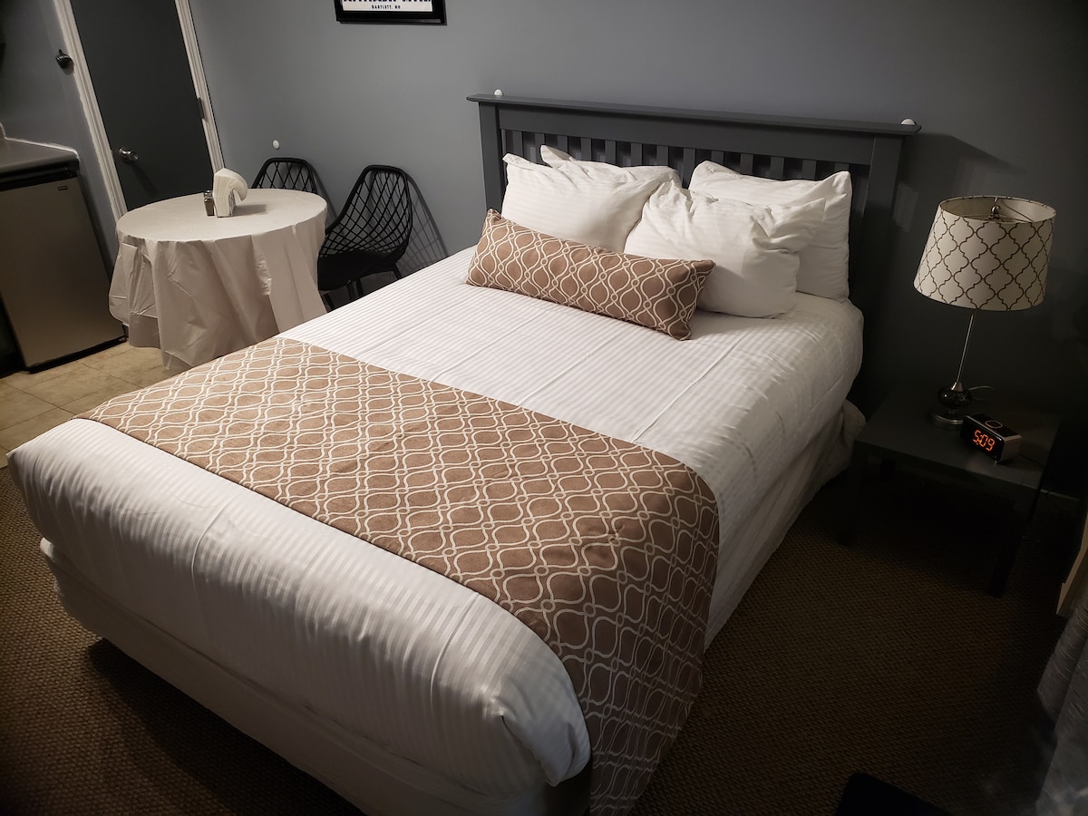 A comfortably furnished sleeping area features a full-size bed with neutral bedding and a decorative throw pillow. A small side table displays a lamp, and a dining table with chairs is positioned nearby, all against a backdrop of muted gray walls.