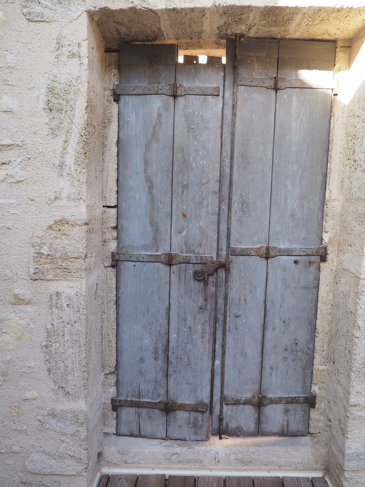 A rustic, weathered wooden door with a muted blue hue is set within a stone wall. The door features sturdy, wrought iron hardware, including hinges and a latch, exemplifying craftsmanship. Sunlight filters through the small opening at the top, casting gentle shadows on the surrounding stone.