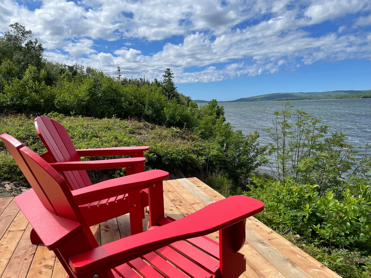 Two red Adirondack chairs are positioned on a wooden deck overlooking a lake. Lush green foliage surrounds the area, with a clear blue sky and scattered clouds in the background, offering a tranquil lakeside view.