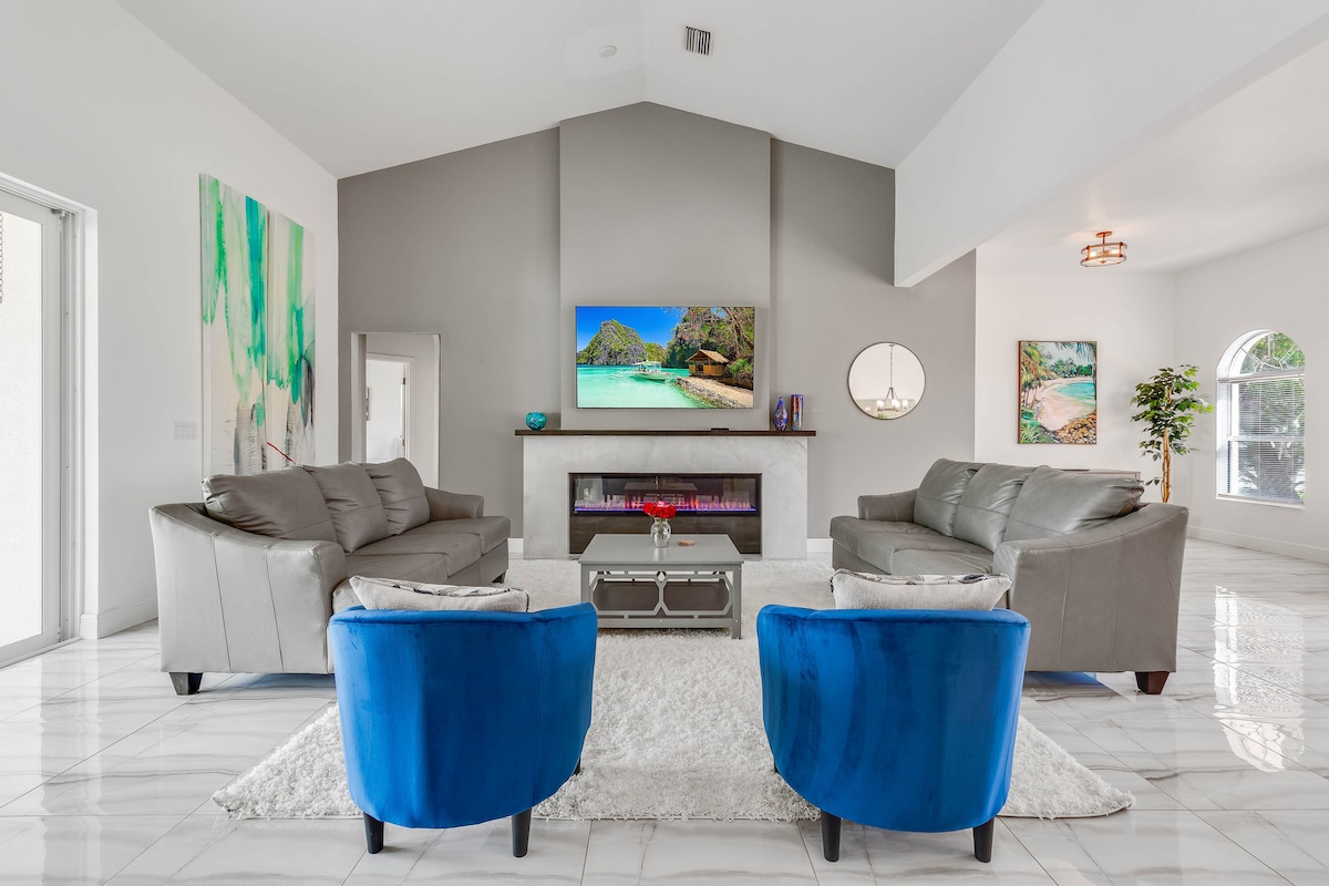 A spacious living area showcases contemporary furnishings, including two gray sofas and two blue accent chairs. A modern fireplace is central, complemented by a large flat-screen TV above it. Natural light enters through large windows, enhancing the inviting atmosphere.